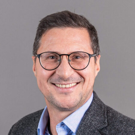 Een man met kort donker haar, een bril en een grijze baard, lacht. Hij draagt een blauw shirt en een donkergrijs jasje, en staat tegen een effen grijze achtergrond.