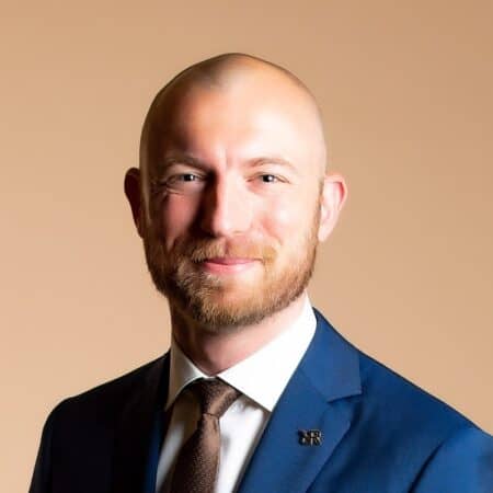 Een kale man met een baard, gekleed in een blauw pak, wit overhemd en bruine stropdas, glimlacht tegen een effen beige achtergrond.
