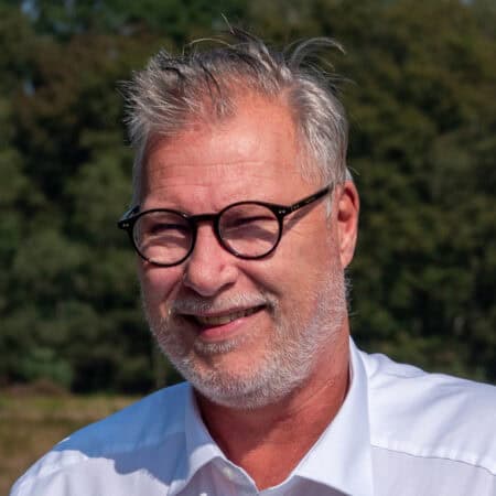 Een persoon met een bril en een wit shirt lacht buiten, met bomen op de achtergrond.