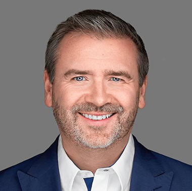 Een man met kort haar en een baard lacht, draagt een wit overhemd en een blauw jasje en staat afgebeeld tegen een grijze achtergrond.