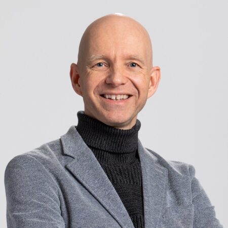Een glimlachende persoon draagt een grijs jasje en een zwarte coltrui, met gekruiste armen, en staat tegen een neutrale achtergrond.