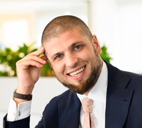 Een man met een baard, gekleed in een pak en stropdas, lacht naar de camera. Hij is binnen met groene planten op de achtergrond.