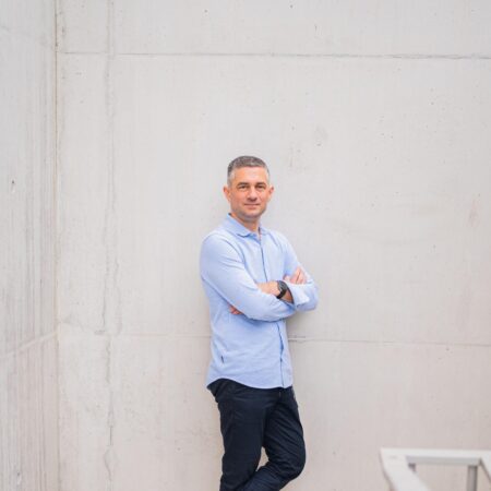 Een persoon in een lichtblauw shirt en donkere broek leunt tegen een betonnen muur met gekruiste armen. Ze dragen witte sneakers en staan op een witte vloer naast een metalen reling.