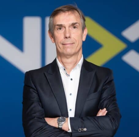 Een man in een donker pak staat met gekruiste armen voor een blauwe achtergrond met witte en groene geometrische vormen.