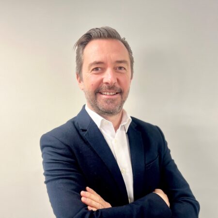 Een man in een donker colbert en een wit overhemd staat tegen een effen, lichte achtergrond, met zijn armen over elkaar, en lacht naar de camera.