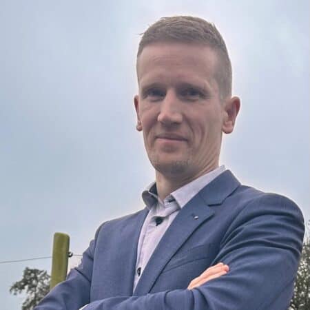 Een man in een blauw colbert en licht overhemd staat buiten met zijn armen over elkaar en kijkt naar de camera. De lucht is bewolkt.
