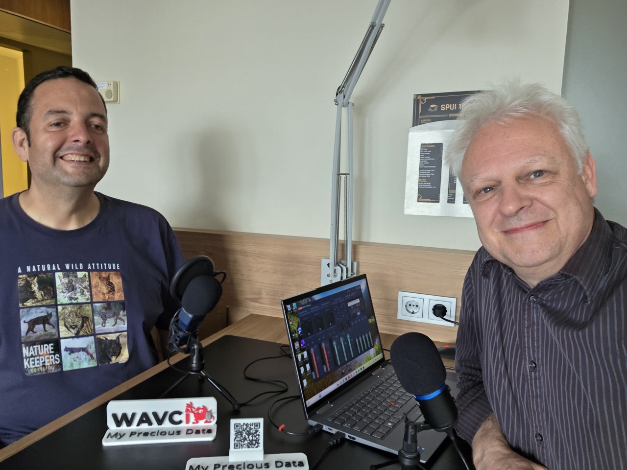 Twee mannen zitten aan een bureau met microfoons en een laptop en nemen een podcast op. De een draagt een grafisch T-shirt, de ander een gestreept shirt. Een WAVC bord is zichtbaar op het bureau.