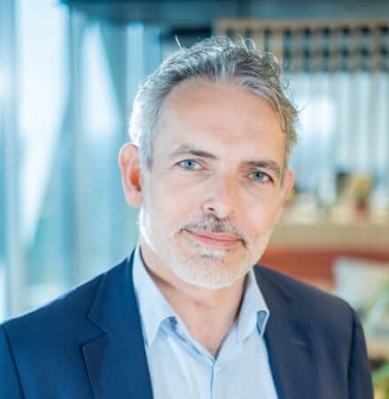 Een man van middelbare leeftijd met grijs haar en een baard, draagt een blauw pak en lichtblauw overhemd, poseert binnenshuis met een onscherpe kantoorachtergrond.