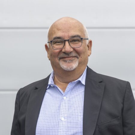 Een oudere man met een bril en een netjes getrimde baard staat tegen een effen lichte achtergrond en draagt een geruit overhemd en een donkere blazer.
