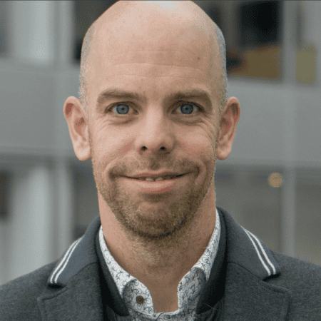 Een man met een kaal hoofd en blauwe ogen met een overhemd met patroon, een groene trui en een jasje staat binnen, kijkt naar de camera en glimlacht lichtjes.