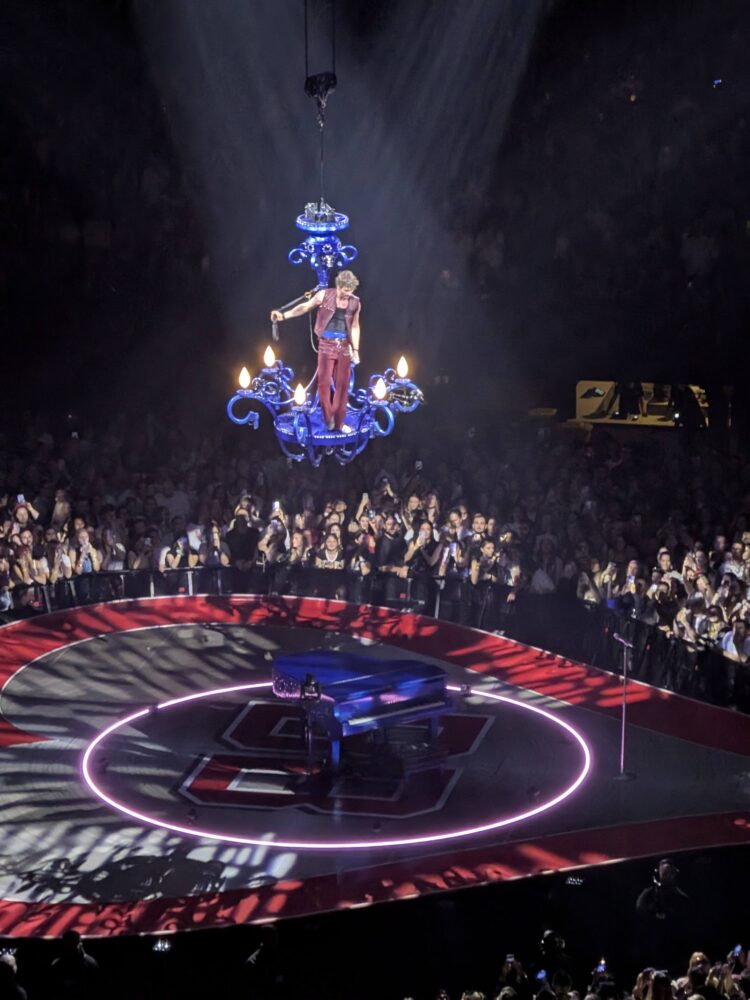Een artiest in een roze broek en een zilveren topje zingt terwijl hij aan een grote kroonluchter boven een cirkelvormig podium hangt terwijl het publiek toekijkt.
