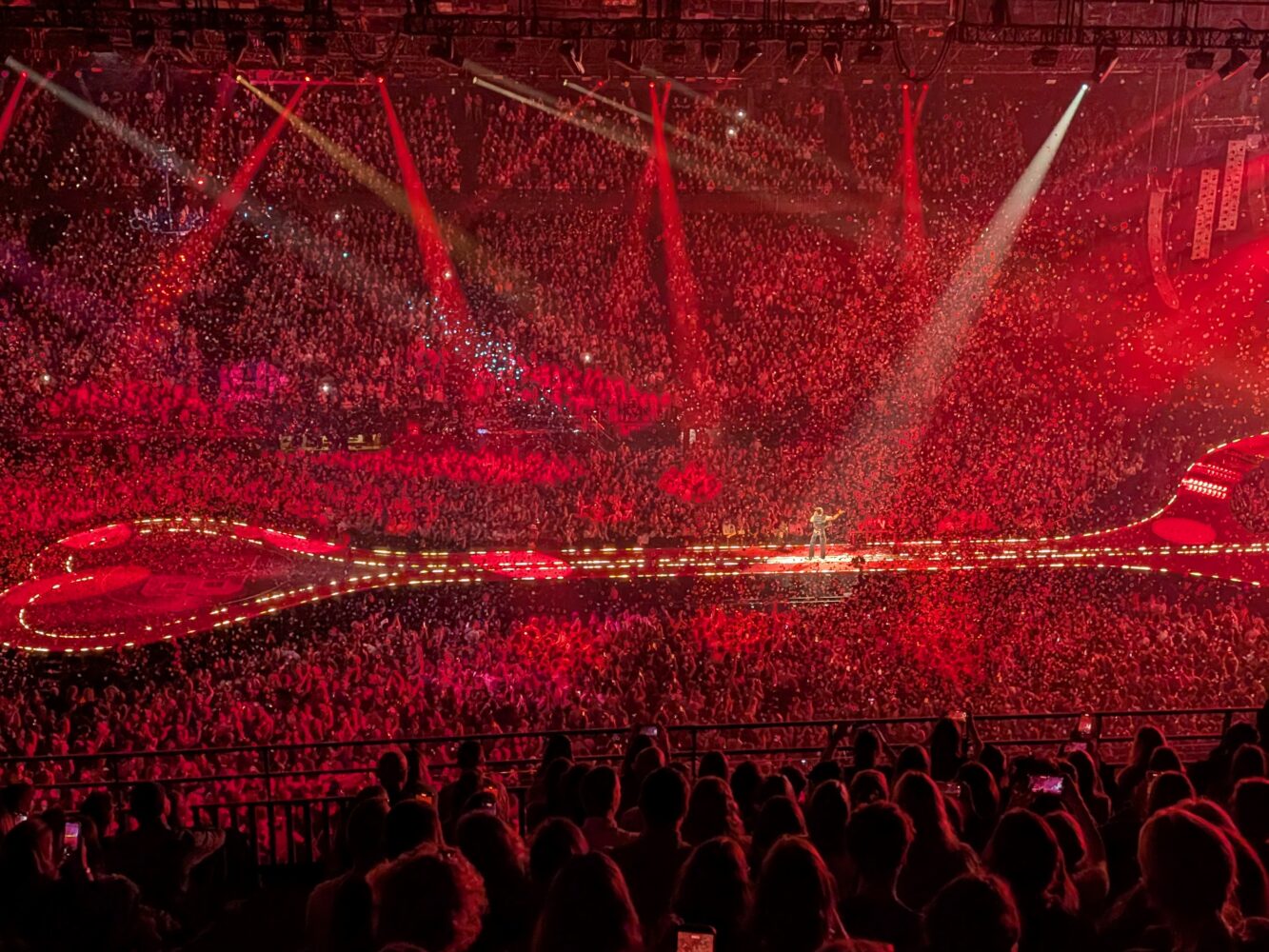 Een grote arena gevuld met een menigte onder rode lichten, met een hartvormig podium en artiest in het midden.