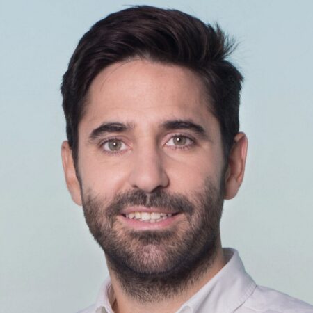 Een man met kort bruin haar, een getrimde baard en een lichtgekleurd overhemd met kraag lacht naar de camera tegen een lichte achtergrond.