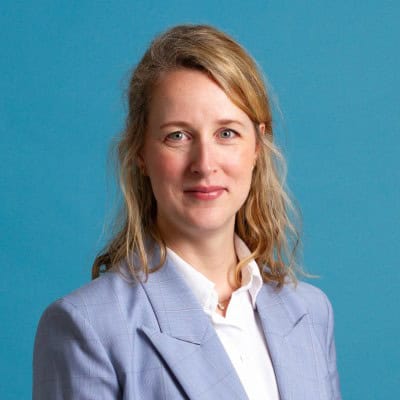 Een vrouw met schouderlang blond haar draagt een lichtblauwe blazer en wit overhemd tegen een effen blauwe achtergrond.