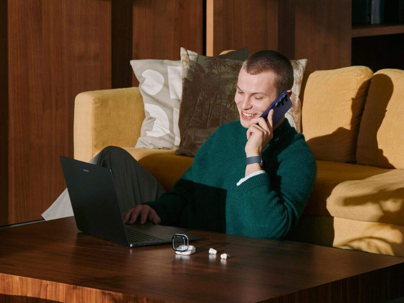 Persoon zit op de grond voor een laptop, praat op een Samsung Galaxy S26 smartphone, met oordopjes en een smartwatch op tafel, in een gezellige woonkamer.