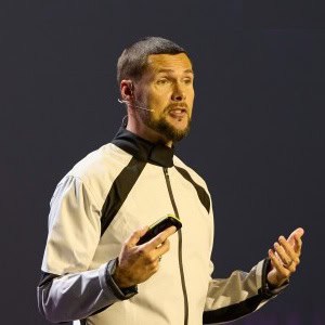 Een man met een korte baard spreekt op het podium, draagt een wit en zwart jasje en een headset-microfoon, en houdt een klein apparaat in één hand.