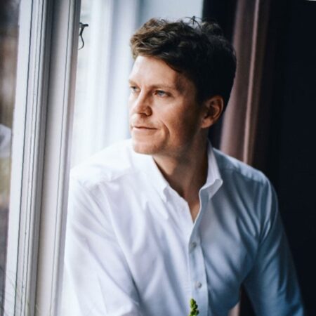 Een man in een wit overhemd kijkt met een bedachtzame uitdrukking uit een raam. Natuurlijk licht verlicht zijn gezicht en op de achtergrond is een gordijn zichtbaar.
