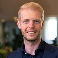 Een man met kort blond haar en een baard, die een donker pak en overhemd draagt, glimlacht naar de camera in een overdekte omgeving.