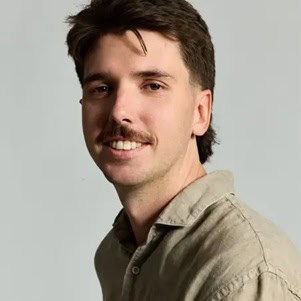 Een man met kort bruin haar en een snor, draagt een beige overhemd en poseert voor een effen lichtgrijze achtergrond.