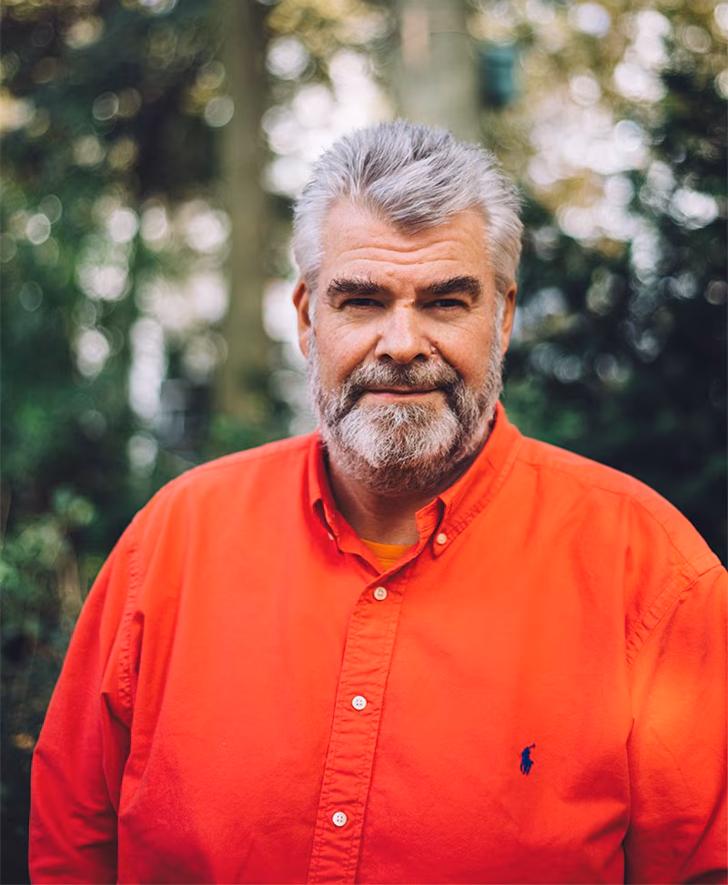 Een man van middelbare leeftijd met grijs haar en een baard draagt een feloranje overhemd met knopen en staat buiten, met bomen op de wazige achtergrond.